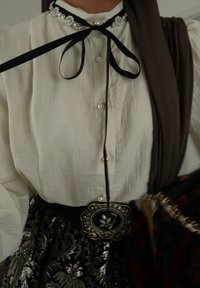 Blouse blanche texturée avec un ruban noir à nouer, ornée de perles ; superposée d'un châle marron. Ceinture noire avec une boucle décorative en or.