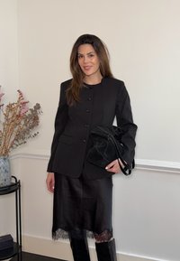 Γυναίκα με μαύρο σακάκι με κουμπιά, φούστα με δαντέλα στο τελείωμα, μπότες μέχρι το γόνατο, κρατώντας μια μαύρη τσάντα, στέκεται σε εσωτερικό χώρο δίπλα σε ένα βάζο με αποξηραμένα λουλούδια.