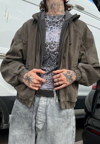Olive roheline bomber-jakk ribitud mansettidega, kantud halli leopardimustriga särgi üle, koos heledate hallide vabalt lõigatud pükstega.