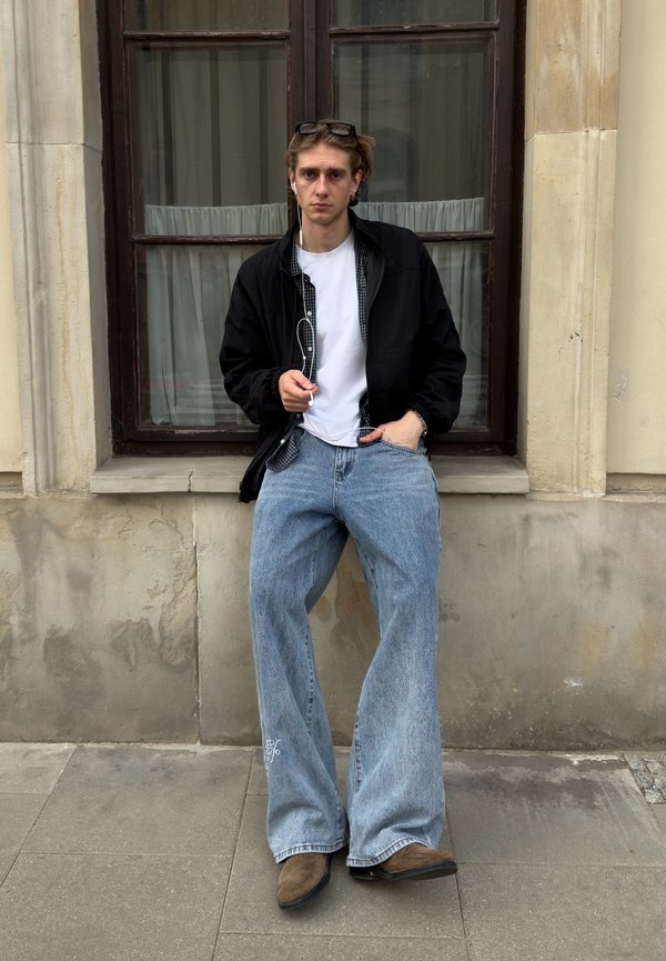 Junge Person trägt schwarze Jacke, weißes Hemd, weite Jeans und braune Schuhe, steht an einer Steinmauer mit Fenster dahinter und hält Kopfhörer.