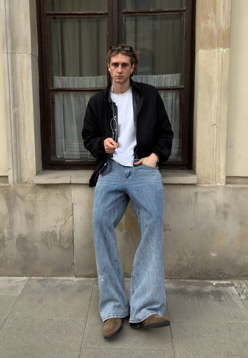 Jeune personne portant une veste noire, une chemise blanche, un jean large et des chaussures marron, debout contre un mur de pierre avec une fenêtre derrière, tenant des écouteurs.