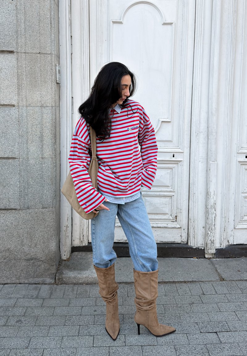 Personne portant un haut à manches longues rayé rouge et gris, un jean bleu clair, des bottes beige décontractées et un sac à bandoulière marron, debout sur le trottoir.