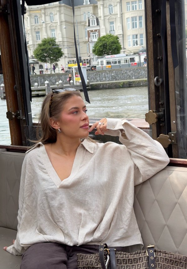 Femme en blouse beige assise sur un banc de bateau au bord de l'eau, regardant vers des immeubles de la ville avec des arbres et des piétons en arrière-plan.