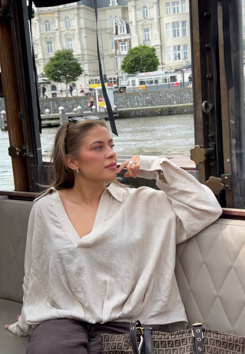 Mujer con blusa beige sentada en un banco de barco junto al agua, mirando hacia edificios de la ciudad con árboles y peatones al fondo.