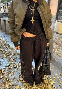 Olivovo zelená bomber bunda, čierne priliehavé tričko, čierne tepláky s dizajnom, čierna taška, viacvrstvové zlaté náhrdelníky, špicaté čierne topánky.