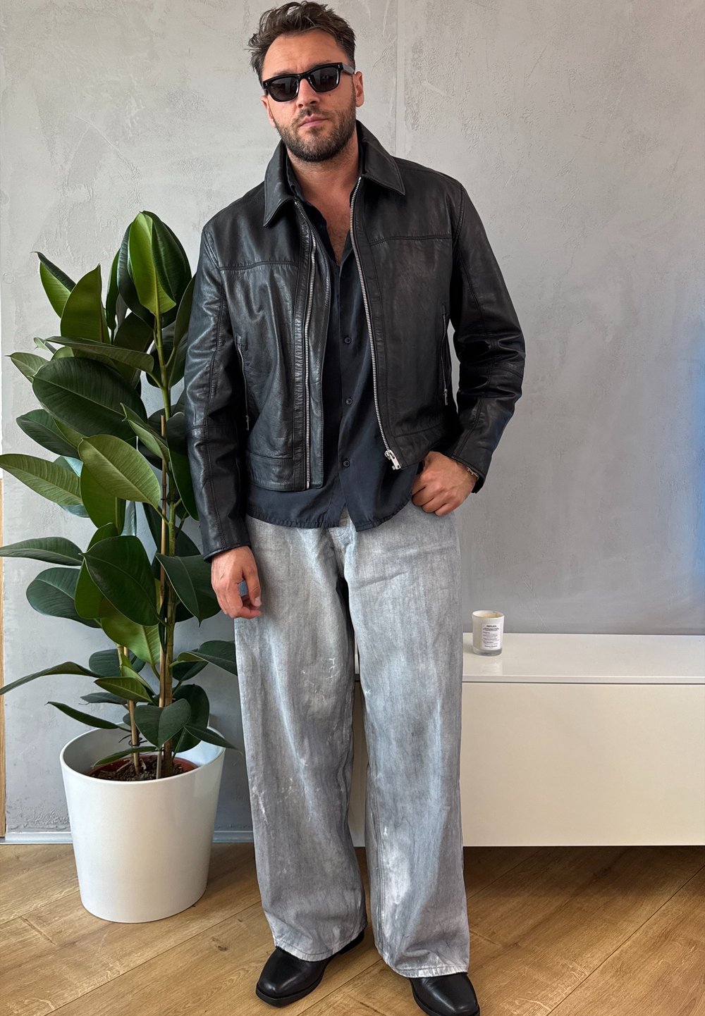 Giacca di pelle nera, camicia nera abbottonata, pantaloni larghi grigio chiaro e scarpe nere. Ambientazione interna con una pianta e una candela.