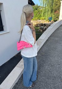 Mulher com cabelo loiro em rabo de cavalo, vestindo uma camisa branca oversized e jeans de perna larga, carregando uma bolsa rosa brilhante, em pé ao ar livre perto de degraus de pedra.