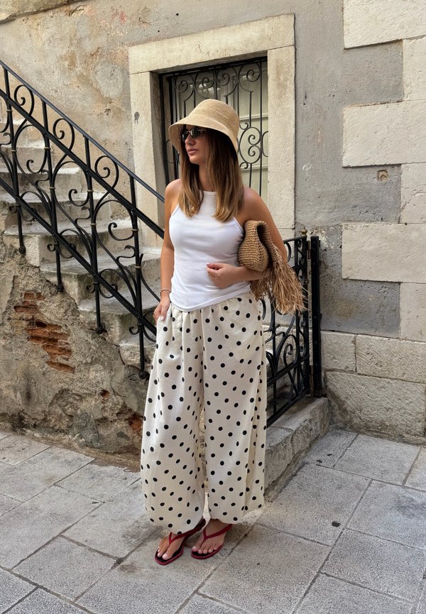 Vrouw met een witte tanktop, brede polkadot-broeken, zonnehoed, zonnebril en rode sandalen staat bij stenen treden met een zwarte ijzeren leuning.