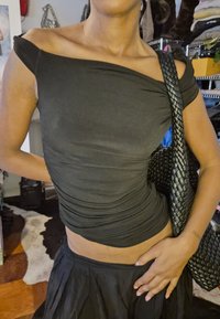 Person wearing a black off-the-shoulder top and skirt, holding a woven black shoulder bag, standing in a room with shelves and clothes.