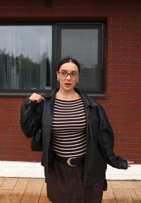 Giacca di pelle nera sopra una maglietta a righe marroni e bianche, cintura nera e gonna marrone. Occhiali rotondi e gioielli d'argento completano il look.