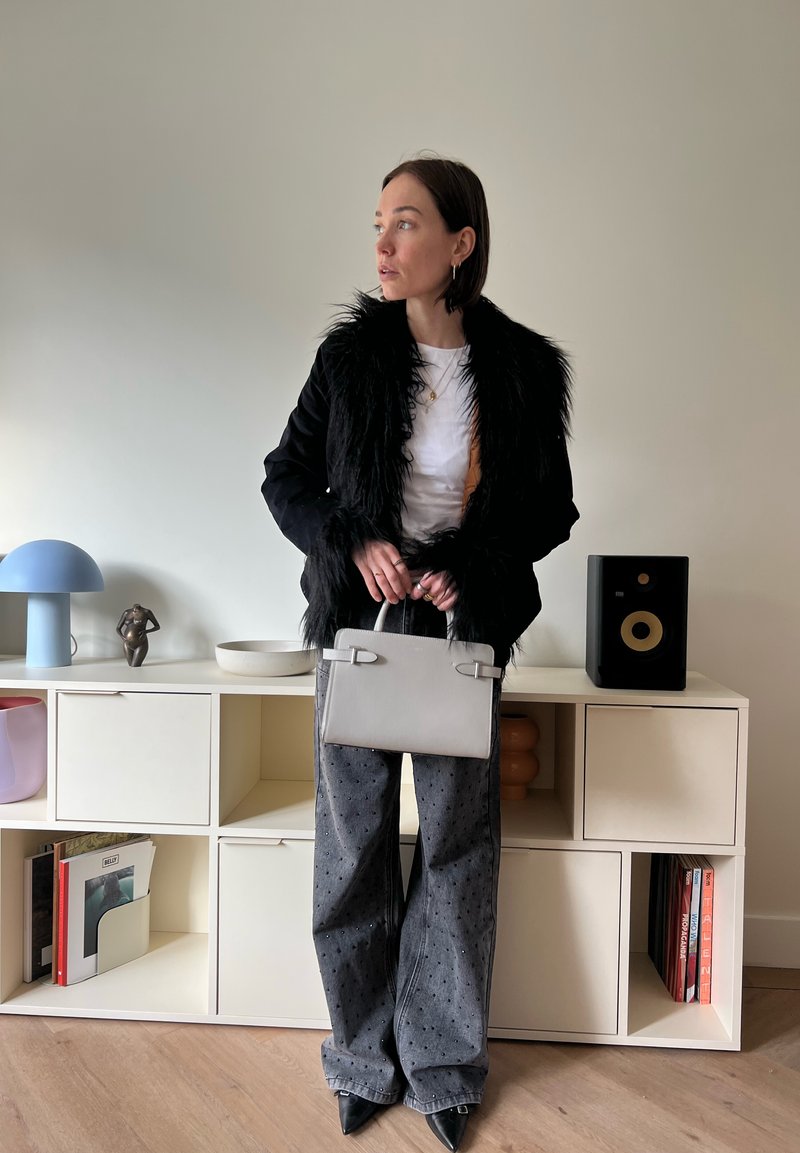 Femme portant une veste noire bordée de fourrure et un pantalon large gris, tenant un sac à main gris clair, debout devant une unité de rangement blanche avec des décorations et des livres.