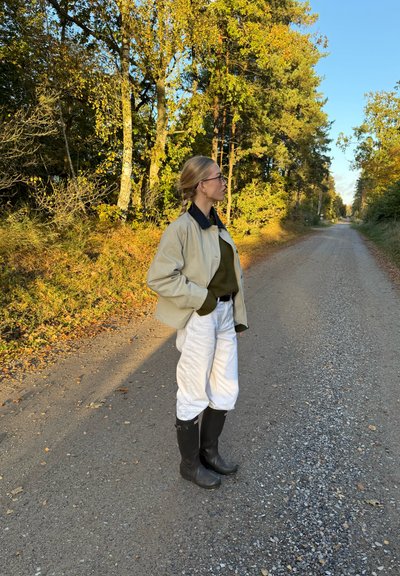 Chaqueta oversized beige, suéter verde oscuro, pantalones blancos y botas de goma negras. El fondo presenta un camino de grava y follaje otoñal.