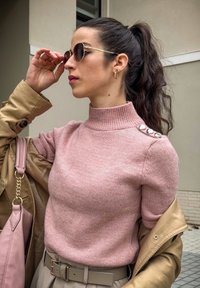 Suéter rosa de malha com um colarinho alto e detalhes em botão nos ombros; combinado com um casaco bege e uma bolsa rosa.