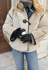 Bézs színű báránybőr kabát fekete szegéllyel és kapoccsal, szürke garbó fölött viselve; kávéscsészét tart, fekete kesztyűt visel.