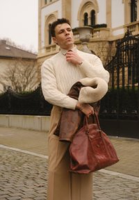Pull en maille crème, veste en cuir doublée de peau lainée marron accrochée au bras, et grand sac en cuir marron. Pantalon beige, cadre urbain.