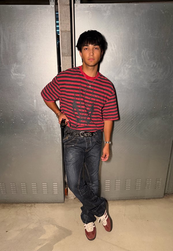 Jeune homme appuyé contre des portes en métal, portant une chemise rayée rouge et noire, un jean foncé et des baskets rouges et blanches, jambes croisées.