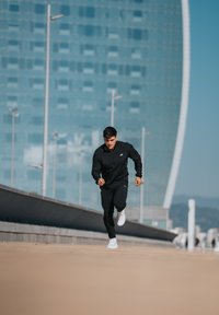 Lekka czarna kurtka i spodnie, białe buty do biegania, mężczyzna biegacz w trakcie kroku na gładkiej powierzchni na tle nowoczesnego szklanego budynku.
