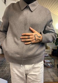 Szürke, cipzáros dzseki gallérral, puha gyapjúszerű anyagból készült. Rejtett oldalzsebekkel rendelkezik. A modell fehér nadrágot visel és tetovált kezeit mutatja.