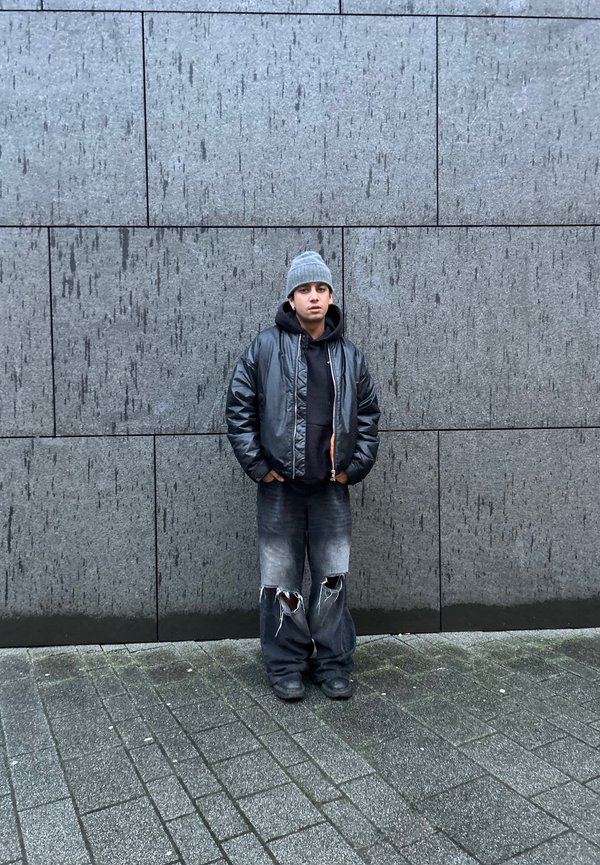 Czarna kurtka bomberka na czarnej bluzie z kapturem, szara czapka beanie i przetarte, oversize'owe czarne dżinsy z postrzępionymi końcami. Stojąc pod szarym murem.
