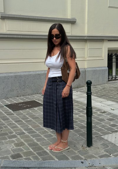 Falda a cuadros negra y gris con detalle de doble hebilla, top de tirantes blanco, bolso de hombro de ante marrón y chanclas ligeras en una calle de adoquines.