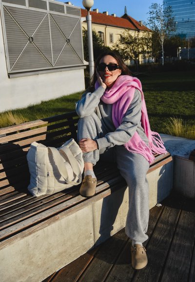 Un chándal gris claro, una bufanda rosa, una bolsa beige con asas y unos zapatos marrones están sobre un banco de madera en un parque verde. Día soleado con edificios de fondo.
