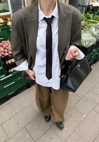 Liela brūna žakete, balta krekla, melna kaklasaiše un brūnas, plašas bikses. Rokā tur melnu ādas rokassomu. Fona attēlā ir pārtikas stendi.