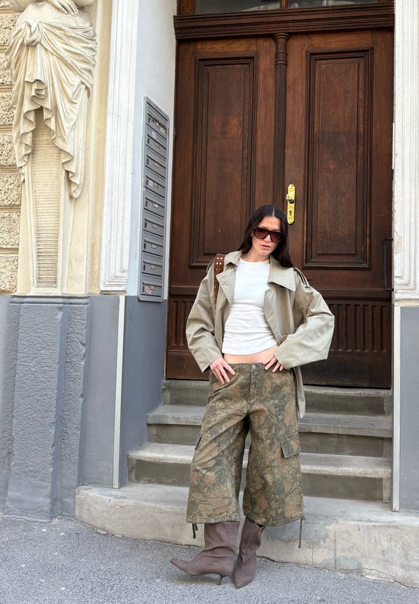 Woman wearing sunglasses, beige trench coat, white top, camo wide pants, and brown boots standing on stone steps in front of a large wooden door.