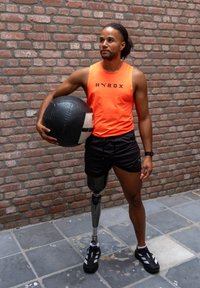 Egy narancssárga ujjatlan felsőt és fekete sortot viselő sportoló fekete-fehér súlylabdát tart a kezében. Próba lábbal és fekete sportcipővel rendelkezik.