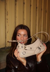 Zapato de charol beige con puntera redonda, correa ajustable, hebillas decorativas y acentos metálicos, sostenido frente a una figura.