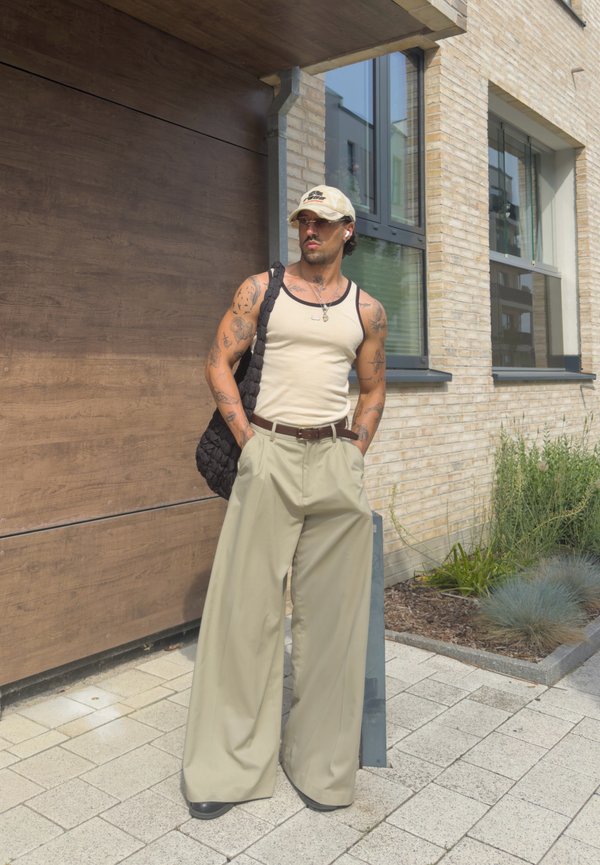 Homme avec des tatouages, portant un débardeur beige, un pantalon large beige, une casquette, des lunettes de soleil et portant un sac matelassé noir, debout à l'extérieur près d'un bâtiment.