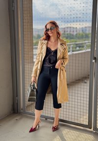 Beige Trenchcoat über einem schwarzen Spitzenoberteil, schwarze Caprihosen und burgunderfarbene Absatzschuhe. Die Accessoires umfassen schwarze Sonnenbrillen und eine gemusterte Handtasche.