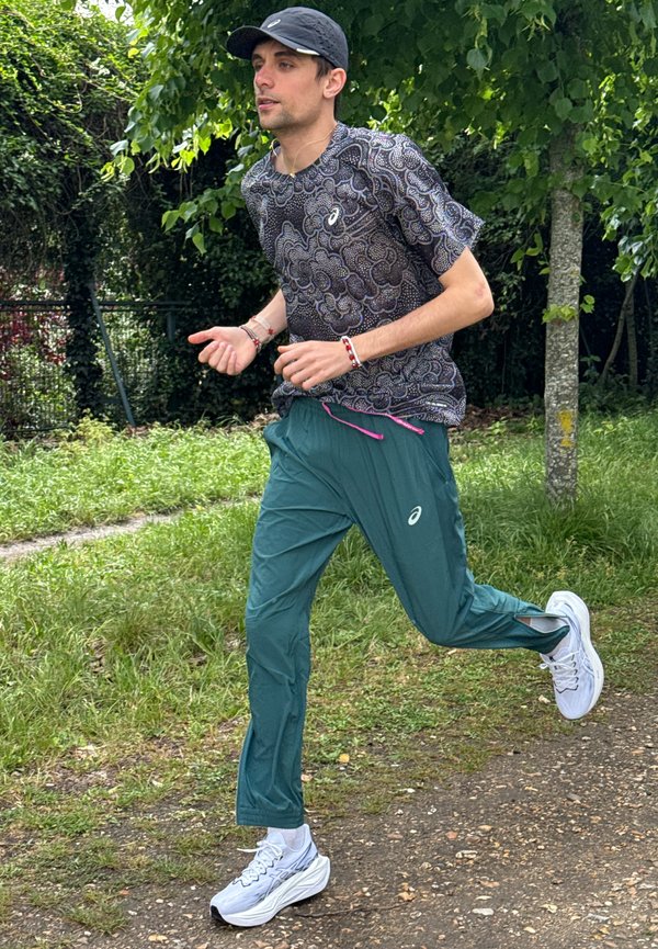 Ein Mann joggt auf einem Naturweg, trägt ein gemustertes Hemd, grüne Hosen, weiße Laufschuhe und eine schwarze Kappe in einem Park.