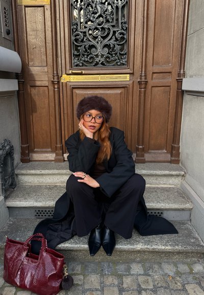 Abrigo negro y pantalones de cintura alta, top de punto marrón, sombrero de piel. Bolso burdeos texturizado con un charm, botines negros, escalones de piedra, puerta de madera.