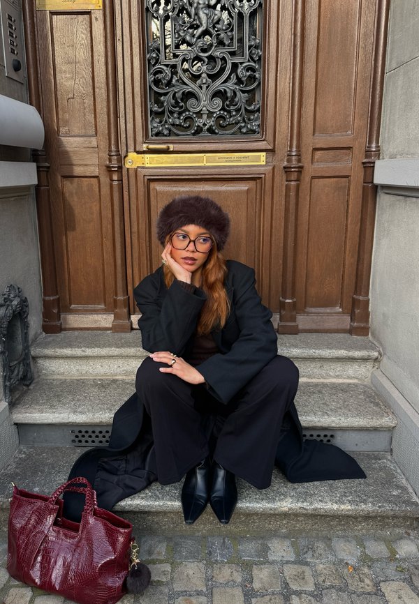 Svart kappa och högmidjade byxor, brun stickad topp, pälsmössa. Vinröd texturerad väska med ett berlock, svarta ankelstövlar, stentrappor, träport.