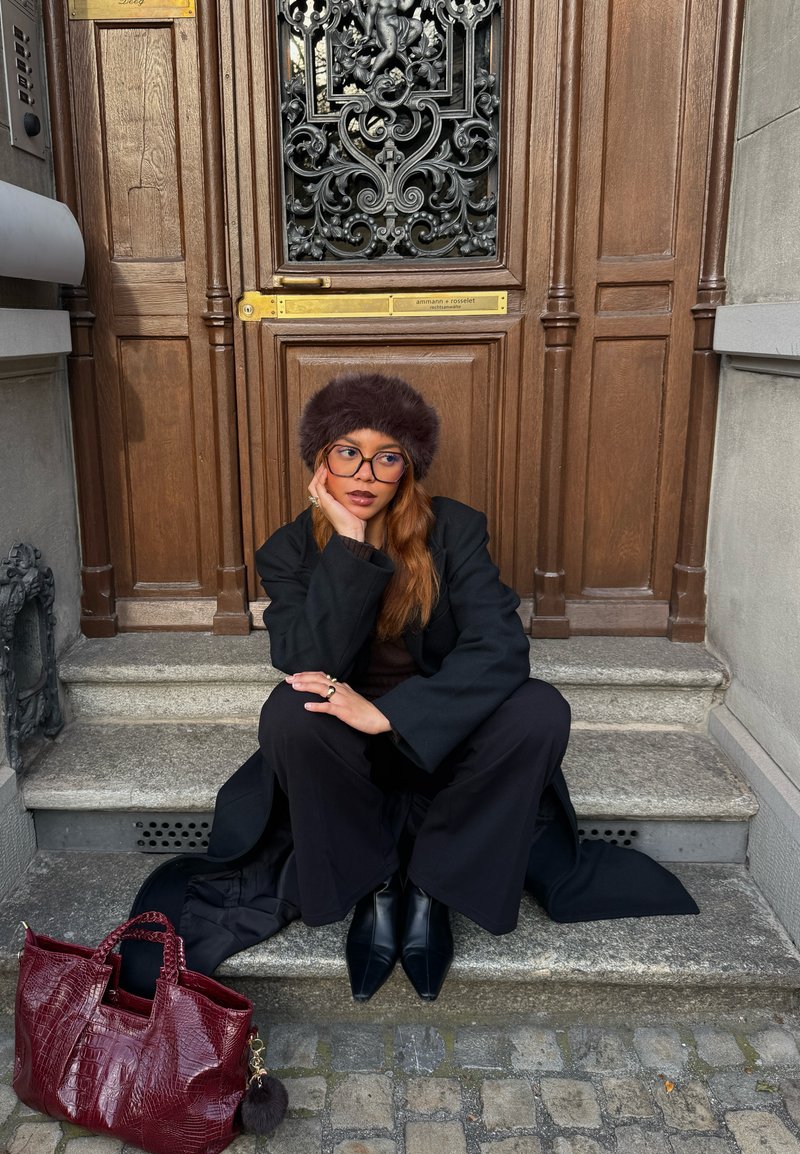 Cappotto nero e pantaloni a vita alta, top marrone lavorato a maglia, cappello di pelliccia. Borsa bordeaux con trama e un ciondolo, stivaletti neri, scalini in pietra, porta in legno.