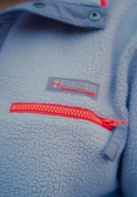 Světle modrý fleecový pulovr s červeným zipovým kapsou a šedou štítkem s nápisem "Columbia Sportswear Company." Měkká textura a neformální design.