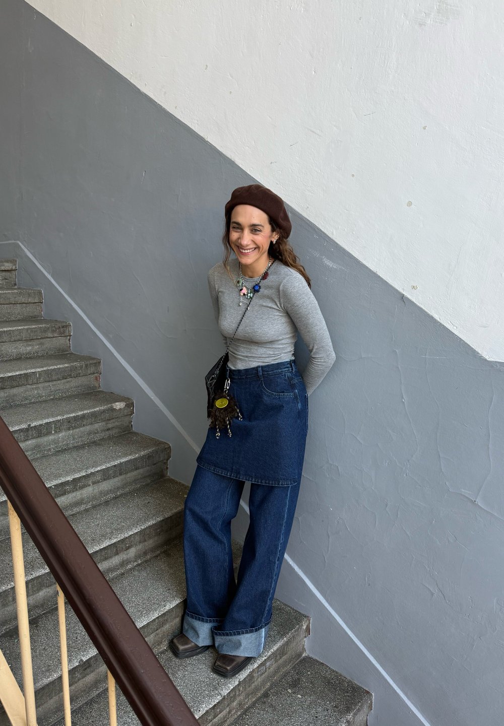 Jupe en denim avec un design de tablier par-dessus un jean à jambes larges, haut à manches longues gris, béret marron et un collier multicolore. Escalier en arrière-plan.