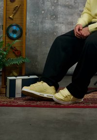Baskets jaunes avec un design perforé et une semelle blanche, assorties à un pantalon noir ample. En arrière-plan, une plante et du matériel audio vintage.