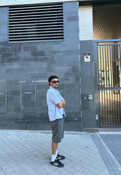 Hombre de pie contra una pared texturizada gris; lleva una camisa de manga corta azul clara, pantalones cortos grises, zapatos negros y gafas de sol.