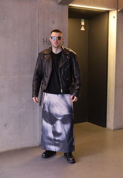 Un modelo se encuentra vestido con una chaqueta de motorista de cuero negro, una camiseta negra y una falda larga con una gran impresión en blanco y negro de un rostro. Lleva gafas de sol reflectantes y botas negras gruesas.