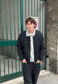 Giacca bomber nera con cerniera frontale, maglione grigio con colletto sotto e jeans scuri. In piedi davanti a un cancello verde.