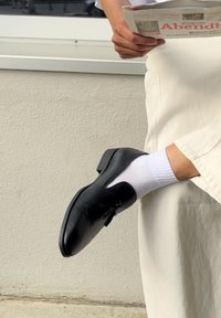 Svart lädersko med spetsig tå och låg klack, i kombination med en vit ribbad strumpa. Skon har en utskärningsdesign.