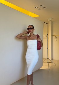 Abito bianco aderente con spalline sottili, abbinato a una borsa rossa texturizzata. Lo sfondo presenta una tonalità neutra e un'illuminazione gialla accentuata.