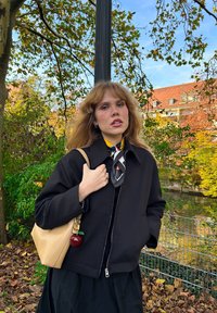 Veste noire zippée sur une écharpe jaune à motifs, associée à une jupe noire et un sac beige clair avec un charm en forme de pomme rouge.