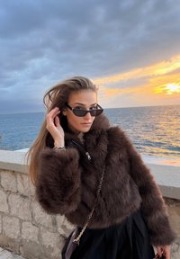 Femme portant des lunettes de soleil noires et une veste en fourrure marron, debout près d'un mur en pierre surplombant la mer lors d'un coucher de soleil sous un ciel nuageux.