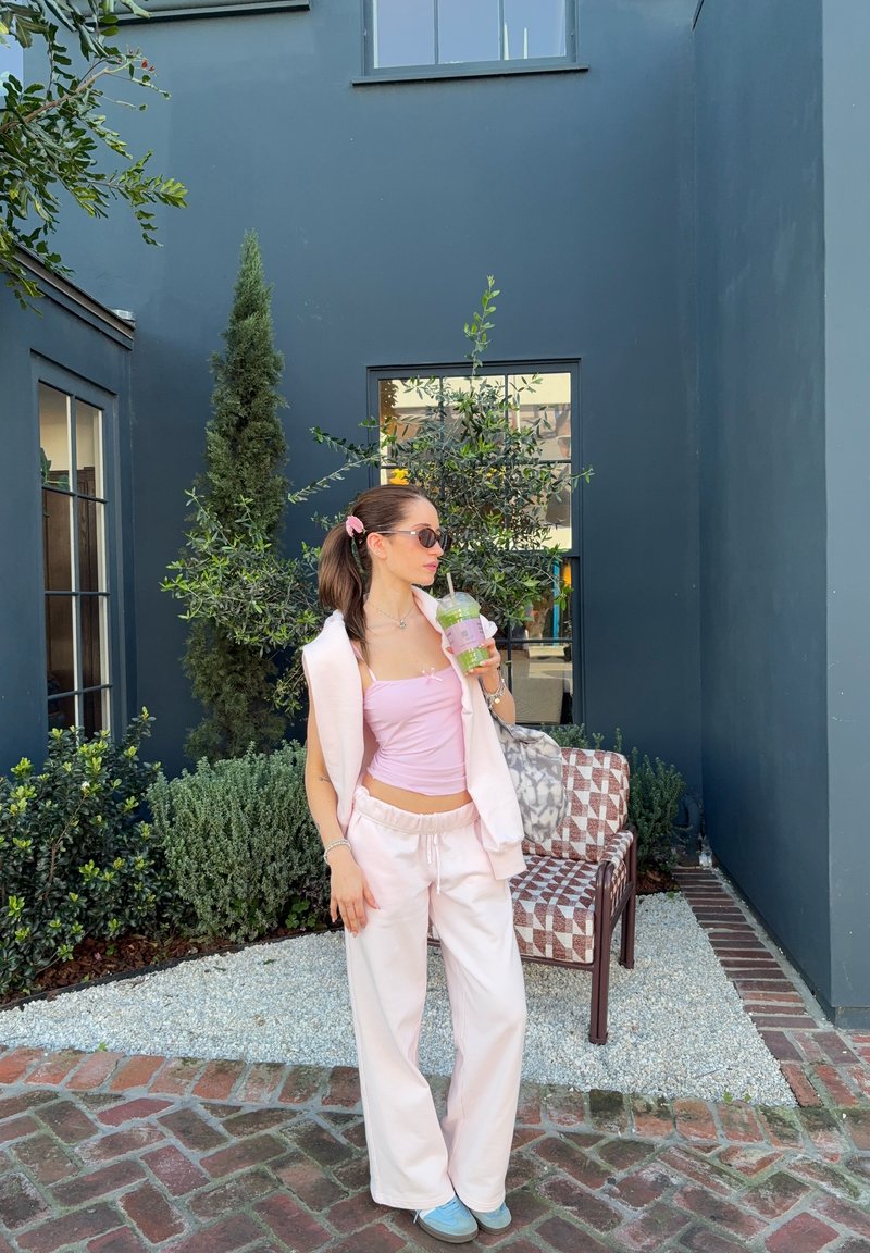 Young woman in pink outfit and sunglasses holds a green drink standing on a brick path near a modern dark blue building and plants.