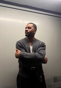 Szürke kötött kardigán bordázott mandzsettákkal, fehér póló fölött viselve. Az egyén magabiztosan áll összefont karokkal, semleges háttér előtt.