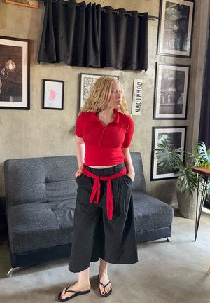 Mujer de cabello claro con camiseta roja y pantalones anchos negros, de pie cerca de un sofá gris en una habitación con cuadros enmarcados y planta de interior.