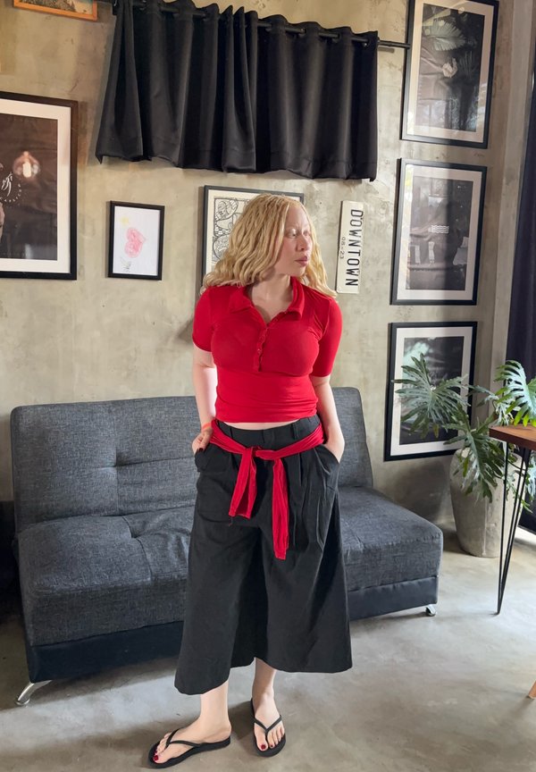 Frau mit hellem Haar in rotem Top und schwarzen weiten Hosen steht neben einem grauen Sofa in einem Raum mit gerahmten Bildern und einer Zimmerpflanze.