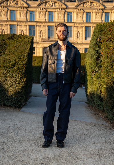 Chaqueta de cuero negra con dos bolsillos frontales, llevada sobre una camiseta de tirantes blanca, combinada con pantalones de mezclilla azul oscuro y zapatos negros, de pie al aire libre.